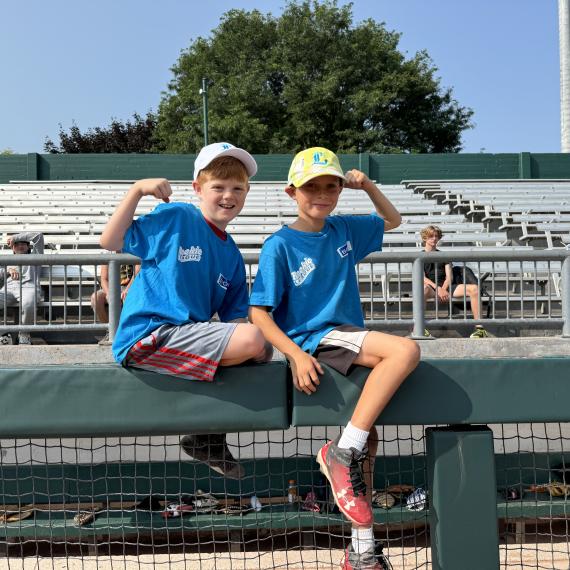Two kids sitting on a fence in a baseball field